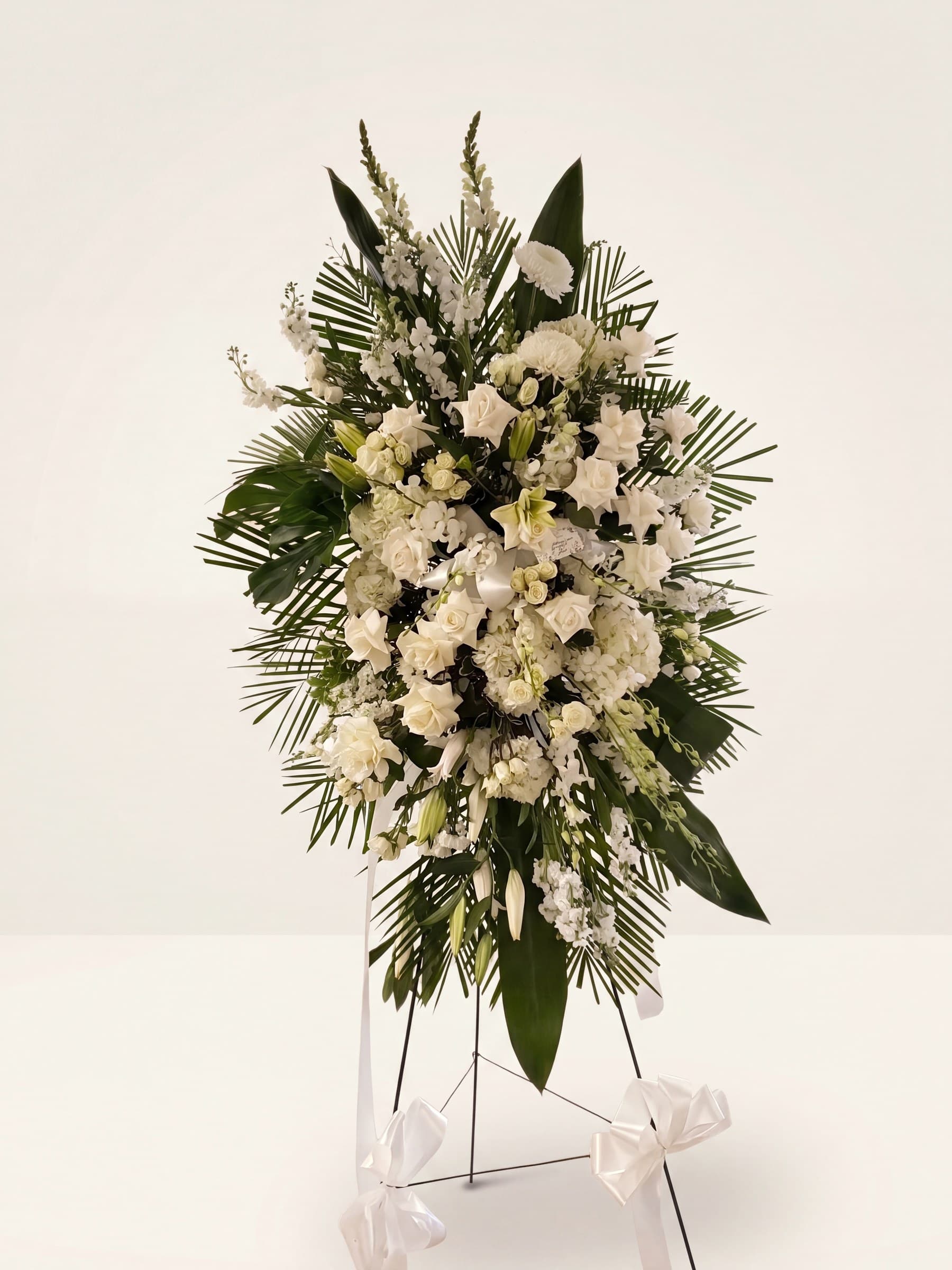 Classic all-white standing funeral spray of roses, snapdragon, and lilies framed in palm on an easel